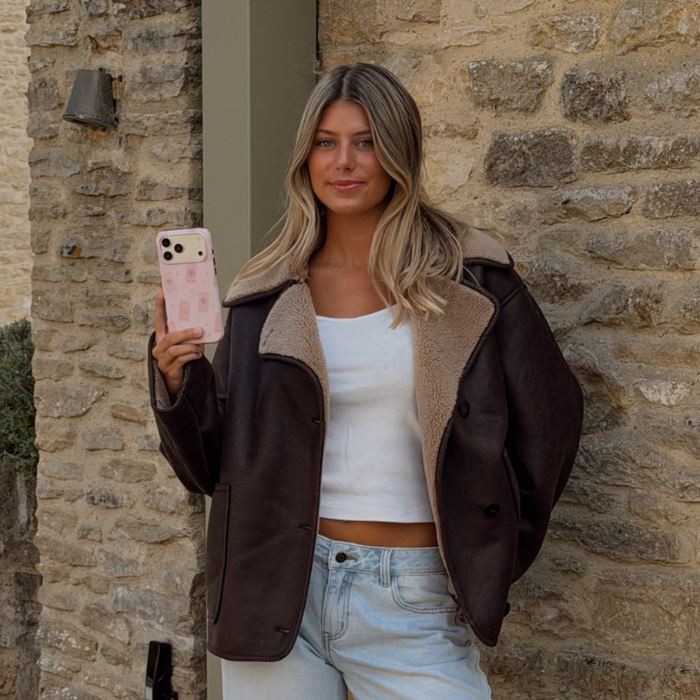 Woman holding a phone up taking a photo, showing a light pale pink phone case with tarot cards and tiny stars. The woman is wearing a brown aviator jacket and light blue jeans.