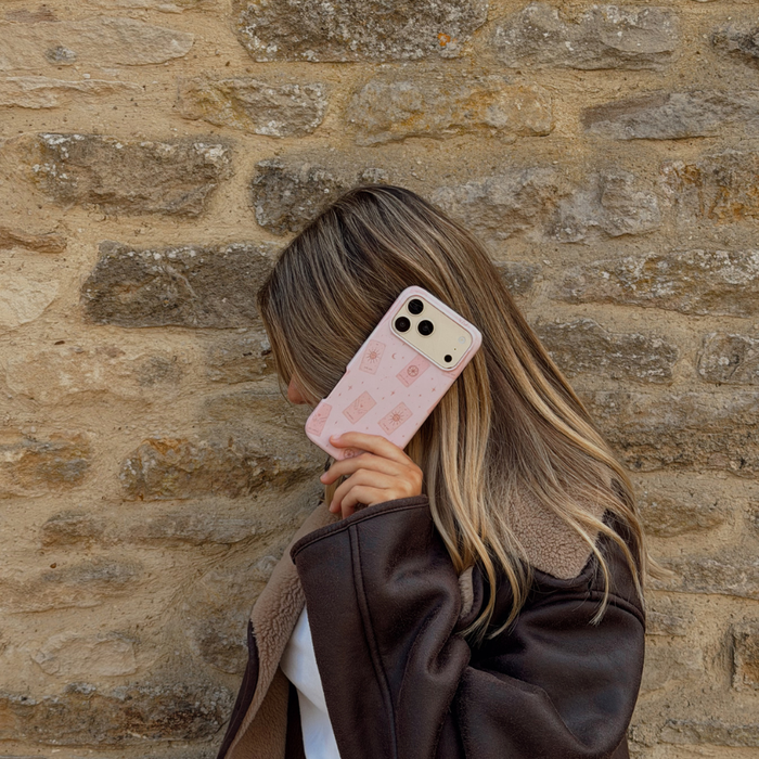 Woman holding a phone up to her ear, showing a light pale pink phone case with tarot cards and tiny stars. The woman is wearing a brown aviator jacket.