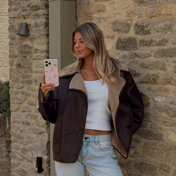 Woman holding a phone up taking a photo, showing a light pale pink phone case with tarot cards and tiny stars. The woman is wearing a brown aviator jacket and light blue jeans.