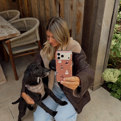 Woman in a brown aviator jacket, holding a phone case with dogs wearing spooky outfits on a brown background, whilst kneeling on the ground stroking a black dog.
