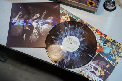A translucent, limited edition Battery vinyl record with a white and blue splatter pattern and its cover—featuring a black-and-white concert crowd photo—rest on a table.