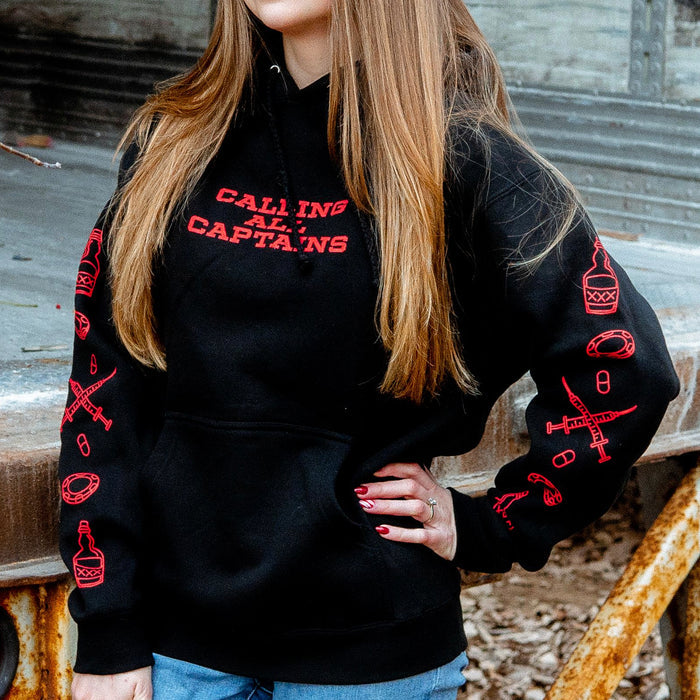 Black pullover hoodie with CALLING ALL CAPTAINS written in red font across the chest. On both sleeves there are drawings of lifesavers, beer, pills, and syringes. An individual is modeling the hoodie and standing in front of a rusted surface.