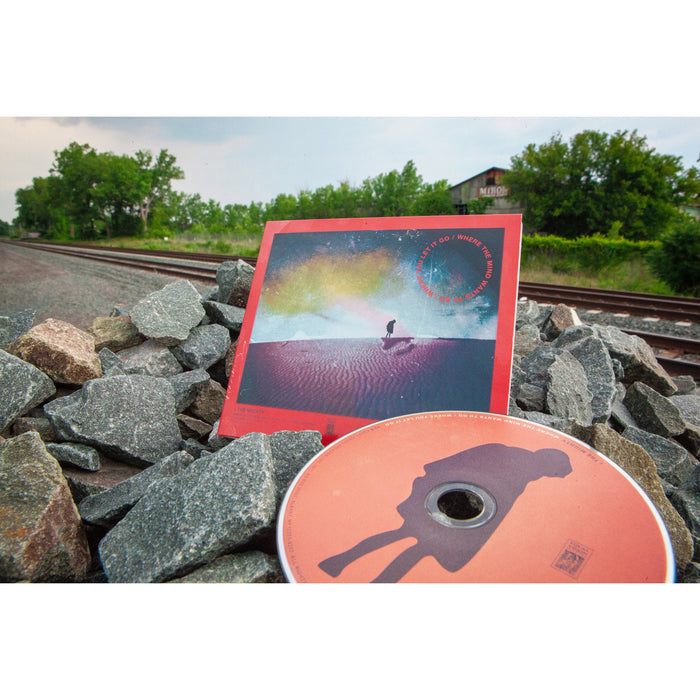 A Let It Go CD by I The Mighty and its album sit on gray rocks by train tracks, with trees and a rustic building behind. The cover shows a silhouette on a red landscape under a colorful sky, referencing Where You Let It Go.