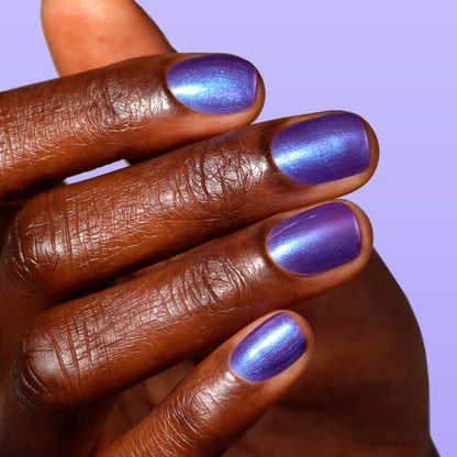A close-up of a hand with dark skin shows short nails painted in LONDONTOWN’s Wildberry Freeze, a metallic polish with a purple-to-blue shimmer, against a light purple background.