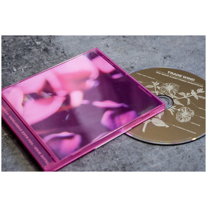 A pink CD case with a blurred floral cover sits on a gray surface next to a gold CD with white floral line art and the track list for Trade Wind’s You Make Everything Disappear.